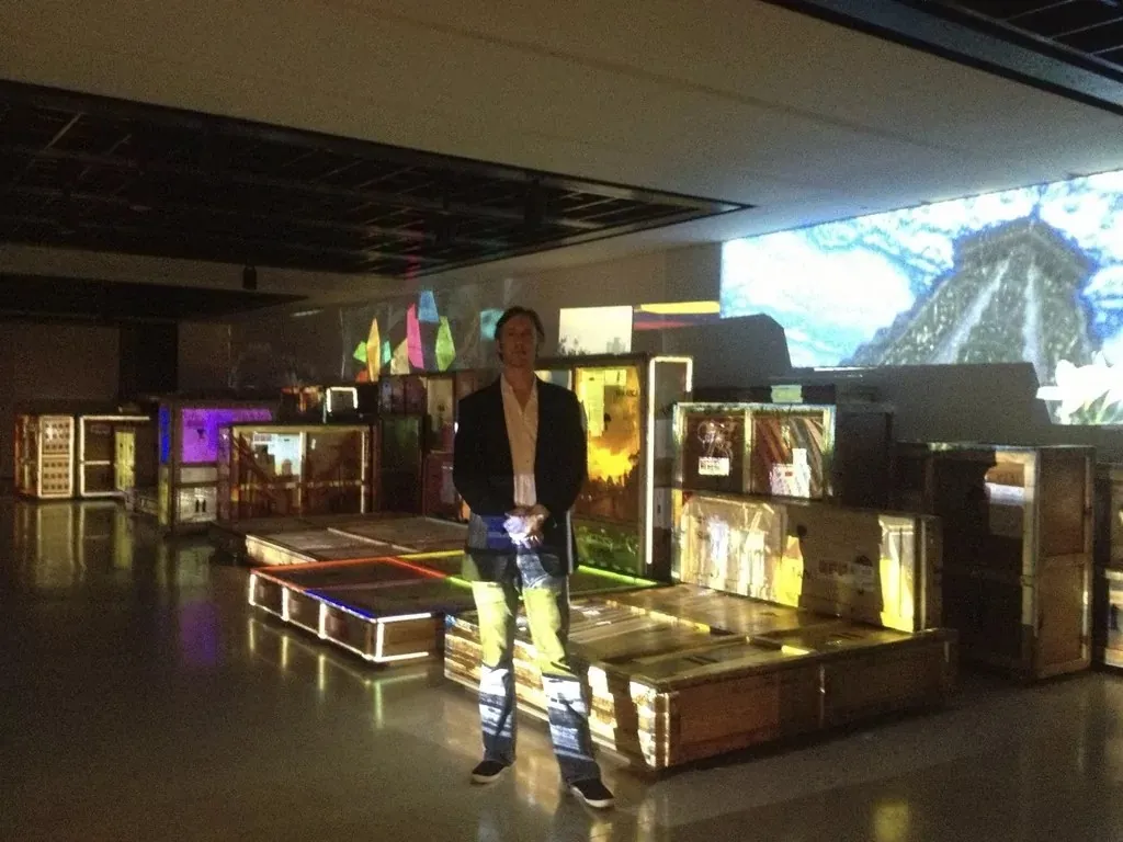 Alex May standing within a darkened installation of projected light and sculptural forms in Caracas