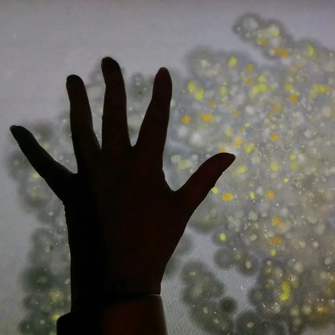 A silhouetted hand reaches across an interactive screen filled with enlarged bacterial colony forms in The Human Super-Organism.