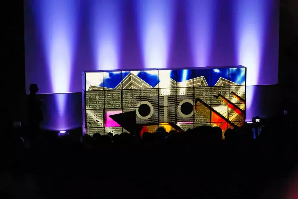 Video-mapped imagery spreads across a wall of illuminated boxes while the audience watches in silhouette.